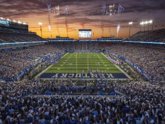 Packed football stadium at sunset with fans, fireworks, and a large scoreboard over the field.