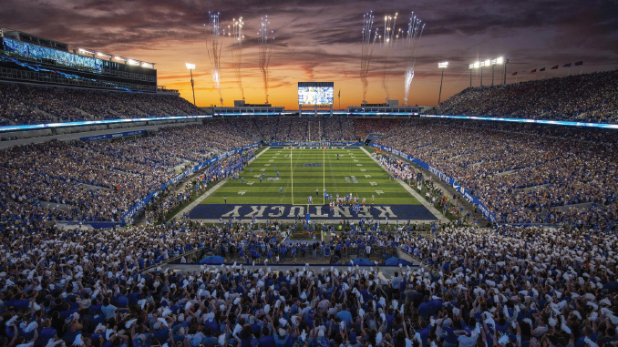 Packed football stadium at sunset with fans, fireworks, and a large scoreboard over the field.