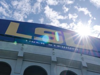 Exterior of a stadium with a large blue wall featuring bold yellow letters and a sun glare over the edge, plus arched entrances below.
