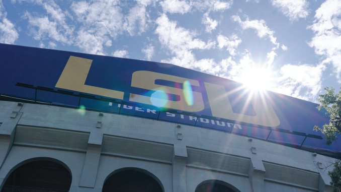 Exterior of a stadium with a large blue wall featuring bold yellow letters and a sun glare over the edge, plus arched entrances below.