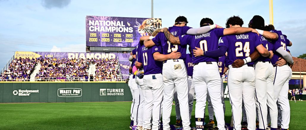 Baseball players in purple uniforms huddle with arms around each other on the field, crowd in purple in the stands behind them.