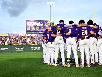 Baseball players in purple uniforms huddle with arms around each other on the field, crowd in purple in the stands behind them.