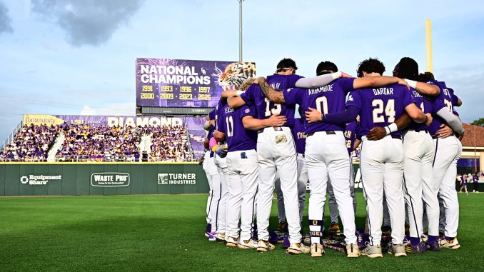 Baseball players in purple uniforms huddle with arms around each other on the field, crowd in purple in the stands behind them.