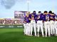 Baseball players in purple uniforms huddle with arms around each other on the field, crowd in purple in the stands behind them.