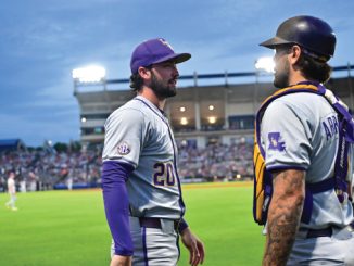 Two gray-and-purple uniformed baseball players stand on the infield and chat, spectators in the stands behind them.