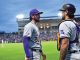 Two gray-and-purple uniformed baseball players stand on the infield and chat, spectators in the stands behind them.