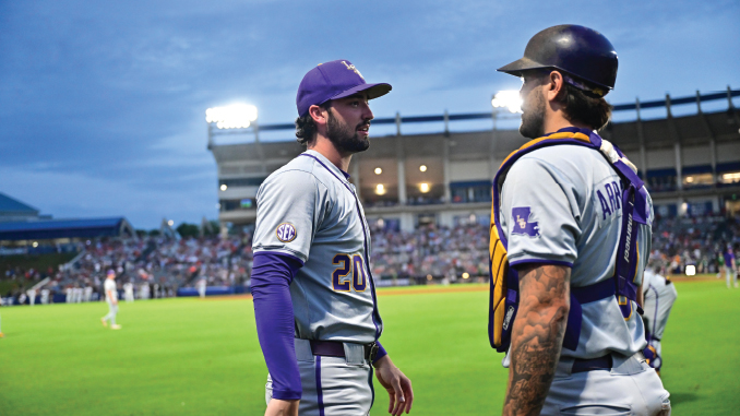 Two gray-and-purple uniformed baseball players stand on the infield and chat, spectators in the stands behind them.