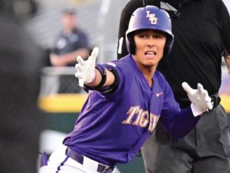 LSU Tigers baseball player in a purple uniform slides toward base with arms outstretched as an umpire watches the play.