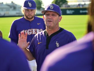 Baseball coach in a purple LSU uniform addressing his players on a sunlit field.