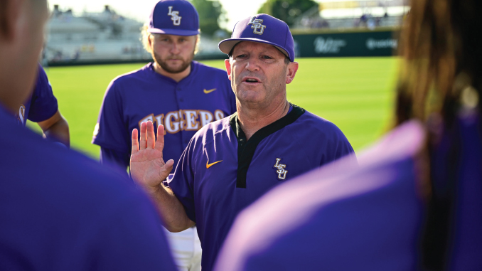 Baseball coach in a purple LSU uniform addressing his players on a sunlit field.