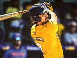 Cricketer wearing a yellow jersey and helmet mid-swing with a bat during a match, looking focused in action.