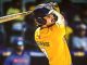 Cricketer wearing a yellow jersey and helmet mid-swing with a bat during a match, looking focused in action.