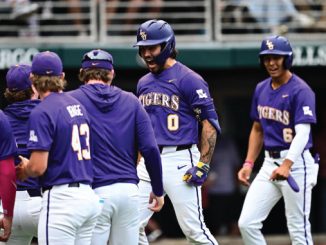 LSU Tigers baseball players in purple and white uniforms stand together in the dugout, celebrating with the center player wearing a helmet (#0).