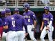 LSU Tigers baseball players in purple and white uniforms stand together in the dugout, celebrating with the center player wearing a helmet (#0).