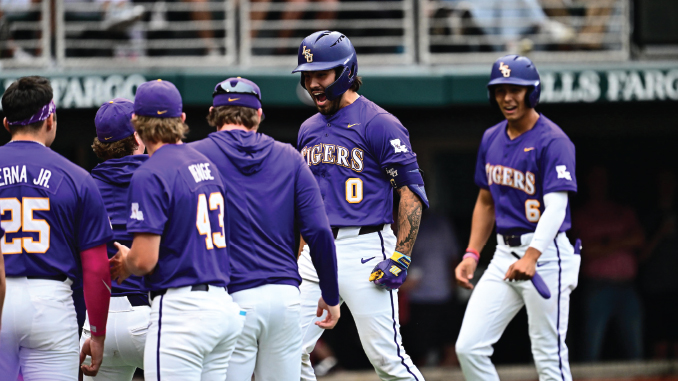 LSU Tigers baseball players in purple and white uniforms stand together in the dugout, celebrating with the center player wearing a helmet (#0).