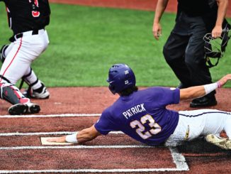 Baseball player Patrick wearing purple with number 23 sliding into home plate while a catcher in black and white reaches to apply tag on a dirt infield with chalk lines visible.