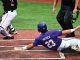 Baseball player Patrick wearing purple with number 23 sliding into home plate while a catcher in black and white reaches to apply tag on a dirt infield with chalk lines visible.