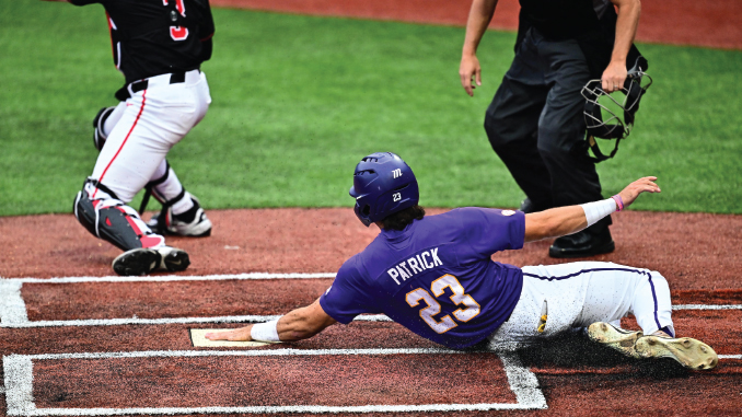 Baseball player Patrick wearing purple with number 23 sliding into home plate while a catcher in black and white reaches to apply tag on a dirt infield with chalk lines visible.