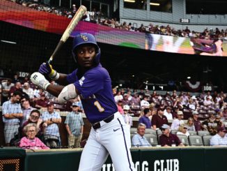 LSU baseball player in a purple uniform swings at a pitch at the plate with a crowd watching from the stands behind the net.
