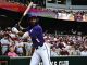 LSU baseball player in a purple uniform swings at a pitch at the plate with a crowd watching from the stands behind the net.