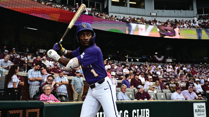 LSU baseball player in a purple uniform swings at a pitch at the plate with a crowd watching from the stands behind the net.