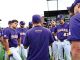 Baseball team in purple uniforms huddles on the field, backs to camera, during a game.
