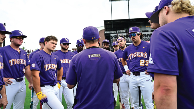Baseball team in purple uniforms huddles on the field, backs to camera, during a game.