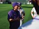 Baseball coach in purple LSU gear talks to players on the field during practice.