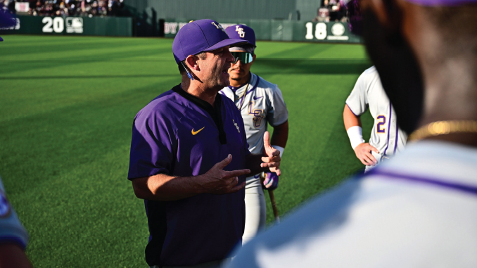 Baseball coach in purple LSU gear talks to players on the field during practice.
