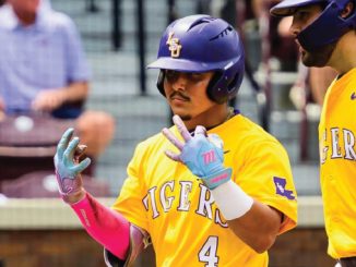 Two LSU Tigers baseball players in yellow jerseys with purple helmets share a high-five on the field; one wears number 4 and pink batting gloves.