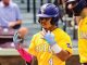 Two LSU Tigers baseball players in yellow jerseys with purple helmets share a high-five on the field; one wears number 4 and pink batting gloves.