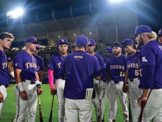 Purple-uniform baseball players form a circle around their coach on the field at night under bright stadium lights, listening intently during a team huddle.