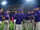 Purple-uniform baseball players form a circle around their coach on the field at night under bright stadium lights, listening intently during a team huddle.