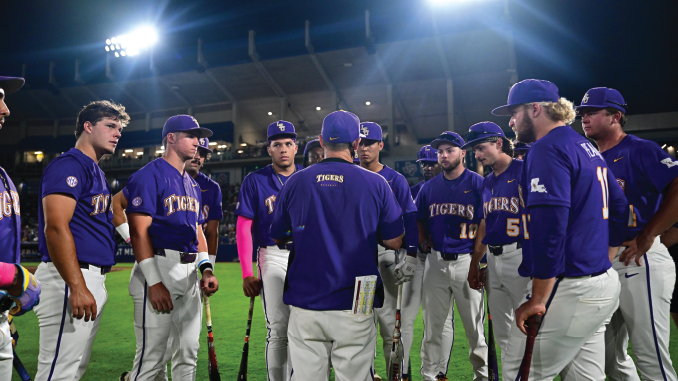 Purple-uniform baseball players form a circle around their coach on the field at night under bright stadium lights, listening intently during a team huddle.