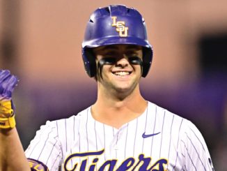 Baseball player in LSU Tigers uniform raises a fist in celebration, wearing a purple helmet with the LSU logo and white pinstriped jersey.