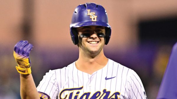 Baseball player in LSU Tigers uniform raises a fist in celebration, wearing a purple helmet with the LSU logo and white pinstriped jersey.