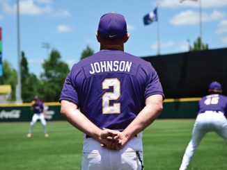 Baseball player Johnson, wearing a purple jersey with number 2, stands with hands clasped behind his back on a sunny practice field as teammates train in the background.