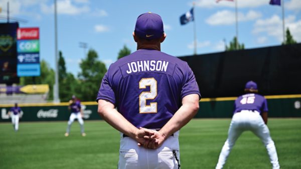 Baseball player Johnson, wearing a purple jersey with number 2, stands with hands clasped behind his back on a sunny practice field as teammates train in the background.
