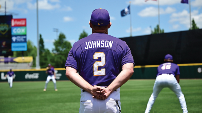 Baseball player Johnson, wearing a purple jersey with number 2, stands with hands clasped behind his back on a sunny practice field as teammates train in the background.