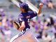 Baseball pitcher in a purple uniform and cap throwing a pitch on the mound with blurred spectators in the stands behind.