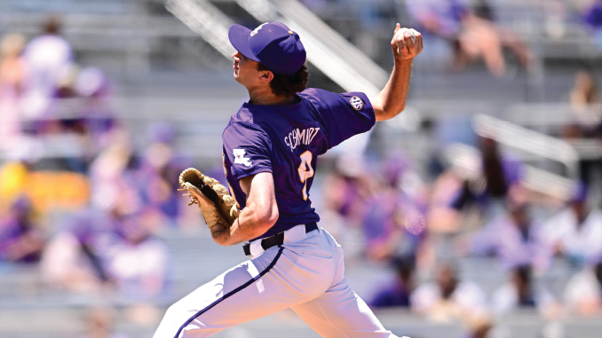 Baseball pitcher in a purple uniform and cap throwing a pitch on the mound with blurred spectators in the stands behind.