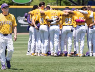 Baseball team in yellow jerseys huddled together on the field; a coach walks in the foreground.
