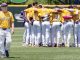 Baseball team in yellow jerseys huddled together on the field; a coach walks in the foreground.