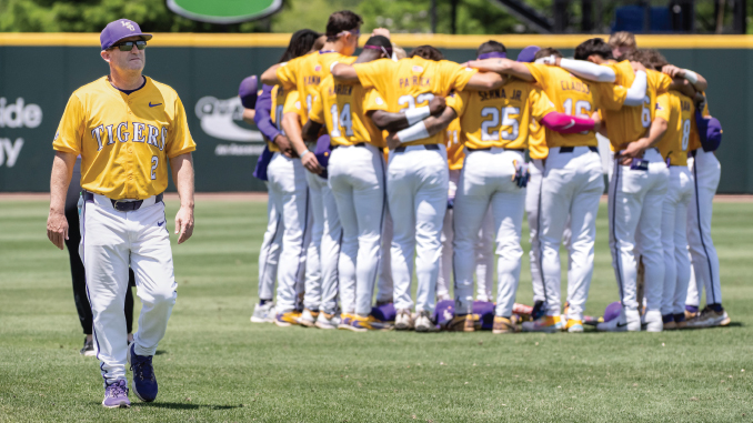 Baseball team in yellow jerseys huddled together on the field; a coach walks in the foreground.