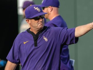 LSU coach in a purple polo shirt and cap with the LSU logo, wearing sunglasses, pointing to the right during practice.