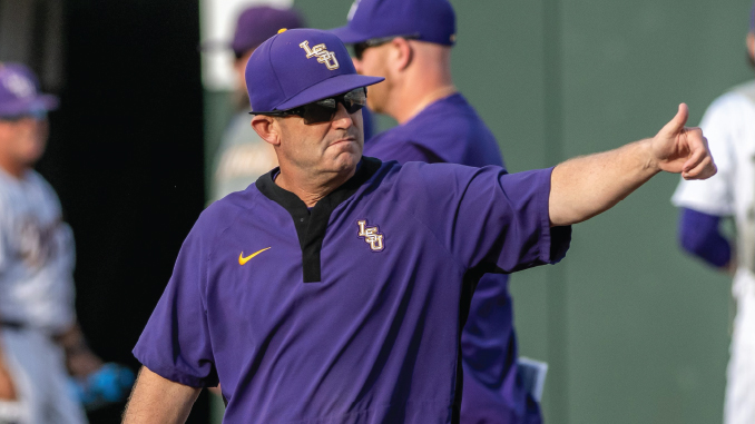 LSU coach in a purple polo shirt and cap with the LSU logo, wearing sunglasses, pointing to the right during practice.