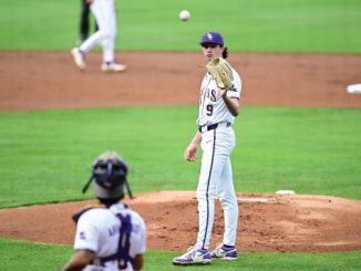 Baseball pitcher on the mound in a white uniform, ball in midair as the catcher crouches in front of home plate.