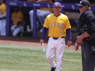 Baseball player in a yellow Tigers uniform with number 2 walks along the field near an umpire, holding a paper.