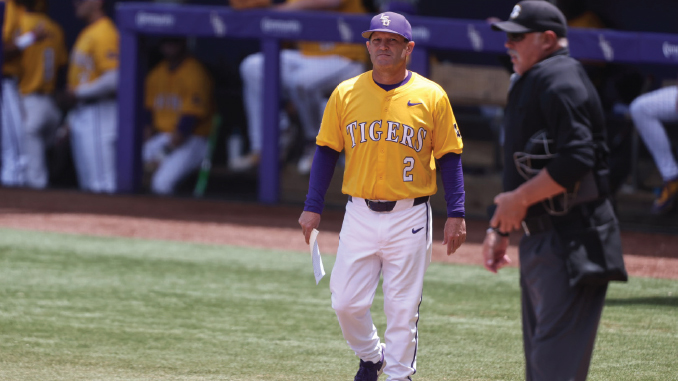 Baseball player in a yellow Tigers uniform with number 2 walks along the field near an umpire, holding a paper.