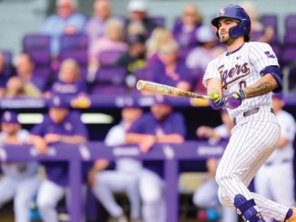 Baseball player in a white pinstripe uniform swings a bat at the plate, helmet on, during a game with a purple-dyed dugout in the background.